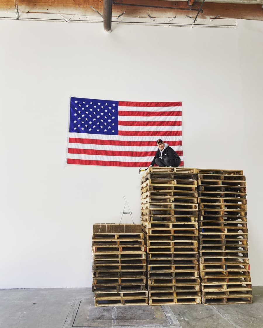 Seal-Bin Han sitting on a stack of pallets in an empty warehouse, a hammer beside him